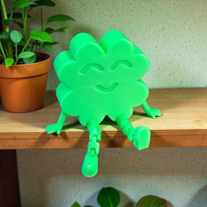 May include: A bright green, smiling, four-leaf clover figurine with articulated legs and arms, sitting on a wooden shelf. A small potted plant is visible in the background, adding a touch of nature to the scene. The clover has a cheerful expression.
