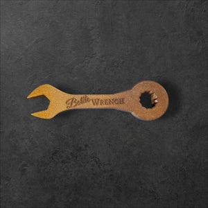 Op de afbeelding: Een bronskleurige flesopener in de vorm van een moersleutel. De tekst "Bottle Wrench" is op het gereedschap gegraveerd. Het heeft een zeshoekig gat aan één uiteinde en een gevorkt uiteinde aan het andere.