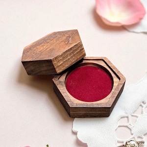 May include: A wooden, hexagonal ring box with a red velvet interior. The box is open, revealing the interior. A diamond ring rests on a white lace cloth. Pink rose petals and a rose are in the background.