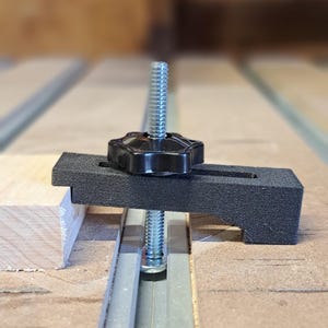 May include: A black hold-down clamp with a threaded rod and a star-shaped knob, designed for woodworking projects. The clamp is positioned on a wooden surface, securing a piece of wood to a track. The background shows an orange machine.