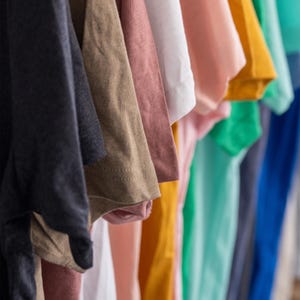 May include: A selection of colourful t-shirts hanging on a rail. The shirts are in a variety of shades, including navy, beige, pink, white, yellow, and turquoise. The image focuses on the sleeves and the fabric's texture.