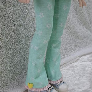 Peut inclure: Un pantalon à pattes d'éléphant vert clair avec un motif floral blanc. Le pantalon a une bordure de dentelle rose en bas et un détail en forme de cœur jaune sur la jambe gauche.