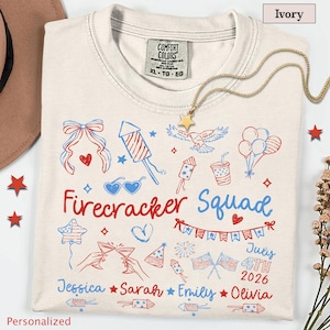 Puede incluir: Una camiseta color marfil con un estampado gráfico en rojo y azul con el texto "Firecracker Squad", "July 4th 2026" y los nombres "Jessica", "Sarah", "Emily" y "Olivia". El diseño incluye ilustraciones de fuegos artificiales, un águila y banderas.