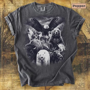 May include: A gray t-shirt featuring a detailed graphic of North American wildlife. The design includes an eagle, bear, wolf, bison, polar bear, and moose. The word "Pepper" is printed on the top right corner of the shirt.