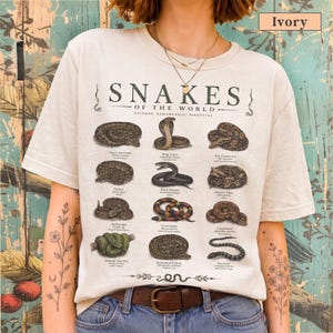 May include: An ivory t-shirt featuring a graphic print titled Snakes of the World with illustrations of twelve different snake species, including an anaconda, king cobra, and boa constrictor, each labelled with its common and scientific name.