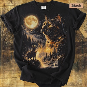 May include: A black t-shirt featuring a graphic print of a large cat head and a smaller cat standing on a rock in a forest landscape under a full moon. The design includes golden light effects, pine trees, and a waterfall. The text Black is visible.