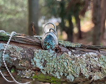 May include: A silver pendant necklace with a turquoise stone. The pendant is oval-shaped with a textured silver border. The necklace chain is a delicate ball chain. The background is a blurred forest scene.