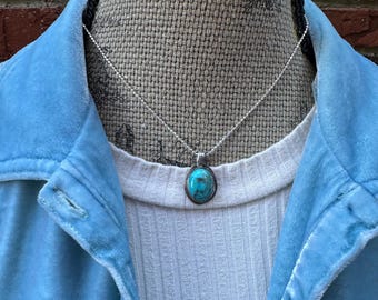 May include: A silver necklace with a turquoise pendant is displayed. The pendant is oval-shaped and set in a silver frame. The necklace is worn over a white ribbed shirt and a light blue jacket.