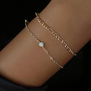 May include: Two gold bracelets are worn on a wrist. One is a delicate chain with a small, round, white gemstone. The other is a thicker chain with an interlocking design. The bracelets are set against a dark background.