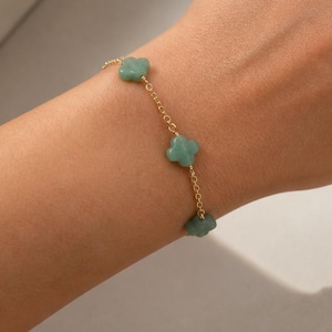 May include: A gold chain bracelet featuring three light green clover-shaped charms. The bracelet is delicate and worn on a wrist. The background is a neutral, off-white color.