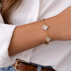 May include: A gold-toned bracelet featuring four-leaf clover charms with white centers and sparkling accents. The bracelet is worn on a wrist, against a white shirt and blue jeans. The bracelet is a delicate piece of jewelry.