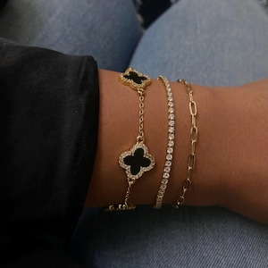 May include: Three gold bracelets. One features a black clover pendant with a gold outline and small clear stones. Another is a tennis bracelet with small clear stones. The third is a gold chain link bracelet.