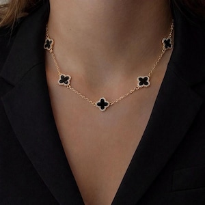 Op de afbeelding: Een goudkleurige ketting met vijf zwarte klavervormige hangers. Elke klaver is omrand met kleine, sprankelende kristallen. De ketting wordt getoond tegen een donkere achtergrond, wat het ontwerp van de sieraden benadrukt.