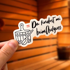 Può includere: Un adesivo bianco con un'illustrazione in bianco e nero di un secchio per sauna e un mestolo. L'adesivo ha il testo "Da findest mi beim Aufguss" in scrittura nera.