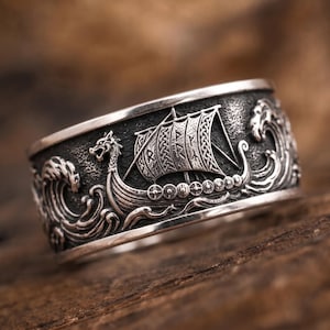 Op de afbeelding: Een brede zilveren ring met een gedetailleerde reliëfsculptuur van een Vikingschip dat door woelige oceaangolven vaart. Het zeil van het schip is versierd met geëtste runensymbolen en de boeg heeft de vorm van een drakenkop.