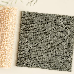 Op de afbeelding: Een beige, cilindrisch object met een rasterpatroon ligt naast een vierkante, grijze, steenachtige tegel. De tegel heeft een oppervlak met textuur dat lijkt op kasseien. De objecten staan op een lichte achtergrond met groene bladsprietjes.