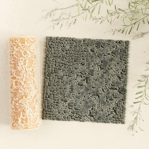 May include: A beige, cylindrical candle with a textured, cracked pattern next to a square, gray stone-like surface. The candle has a rough, organic appearance. The stone-like surface has a cobblestone design. Decorative sprigs are in the background.
