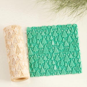 May include: A beige rolling pin and a green textured mat, both decorated with a repeating Christmas tree pattern. The rolling pin is cylindrical, while the mat is rectangular. The trees are white and detailed.