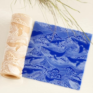 Puede incluir: Un rodillo de amasar beige y una lámina cuadrada azul, ambos con un diseño de ballenas y olas. El rodillo de amasar tiene un patrón repetitivo de ballenas y olas. La lámina azul tiene un patrón detallado de ballenas y olas.