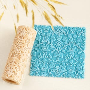 Può includere: Un mattarello beige e un tappetino blu strutturato con un motivo floreale. Il mattarello ha un motivo floreale simile. Il tappetino è di forma quadrata con un motivo ripetuto di fiori e foglie.