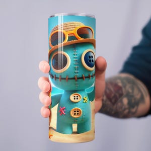 May include: A tall, cylindrical tumbler featuring a cartoon character design. The character is a teal-colored doll with button eyes and a stitched appearance, wearing a straw hat and sunglasses. The tumbler is held by a person.