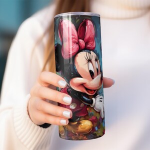 May include: A tall, cylindrical tumbler featuring a colorful illustration of Minnie Mouse. The design includes a large pink bow, a smiling face, and a red dress. The tumbler is held by a person wearing a white sweater.