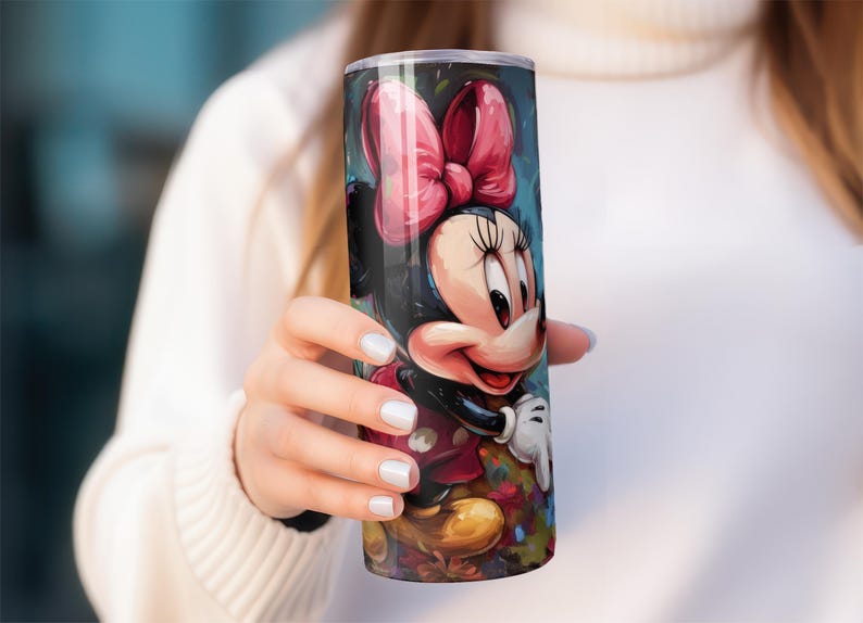 May include: A tall, cylindrical tumbler featuring a colorful illustration of Minnie Mouse. The design includes a large pink bow, a smiling face, and a red dress. The tumbler is held by a person wearing a white sweater.