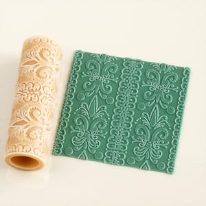 May include: A beige rolling pin and a green textured square. The rolling pin has a repeating floral design. The green square has a repeating floral and lace design. The items are likely for baking or crafting.