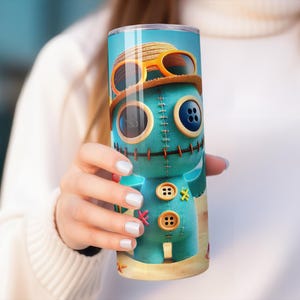 May include: A teal tumbler featuring a cartoon character with button eyes, stitched details, and a straw hat with sunglasses. The design includes a beach scene at the bottom. The tumbler is cylindrical and appears to be made of stainless steel.