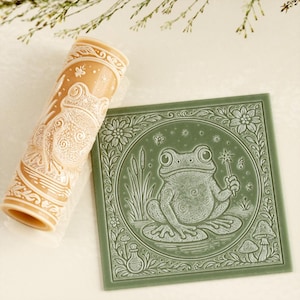 May include: A tan rolling pin and a green square tile with a frog design. The rolling pin has a frog and floral pattern. The tile features a frog holding a wand, surrounded by flowers and plants.