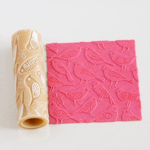 Pode incluir: Um rolo de massa bege e um quadrado rosa com um padrão de pássaros. O rolo de massa tem um desenho de pássaros repetido. O quadrado rosa tem um padrão de pássaros em relevo.