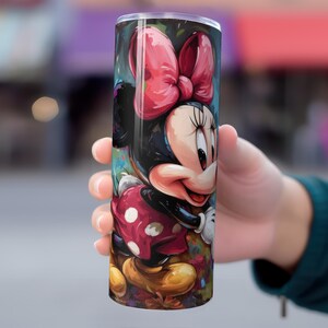 May include: A cylindrical tumbler featuring a colorful illustration of Minnie Mouse. The design showcases Minnie's iconic red bow, polka-dot dress, and smiling face. The tumbler is held in a person's hand.
