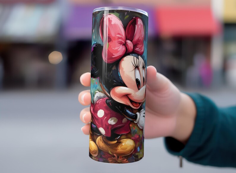 May include: A cylindrical tumbler featuring a colorful illustration of Minnie Mouse. The design showcases Minnie's iconic red bow, polka-dot dress, and smiling face. The tumbler is held in a person's hand.