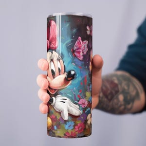 May include: A tall, cylindrical tumbler featuring a colorful illustration of Minnie Mouse. The design includes a pink bow, white gloves, and a vibrant floral and butterfly background. The tumbler is held by a person.