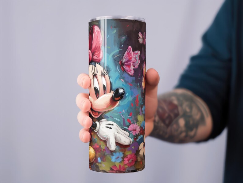 May include: A tall, cylindrical tumbler featuring a colorful illustration of Minnie Mouse. The design includes a pink bow, white gloves, and a vibrant floral and butterfly background. The tumbler is held by a person.
