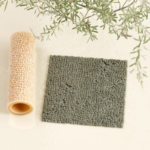 May include: A beige, cylindrical roller with a textured, open-weave pattern, and a square, gray, stone-like tile. The roller is positioned vertically, and the tile is to its right. Greenery is in the background.