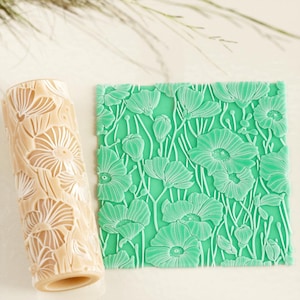 Może przedstawiać: Beżowy wałek do ciasta i zielony kwadrat z kwiatowym wzorem. Wałek do ciasta ma powtarzający się kwiatowy wzór. Zielony kwadrat ma podniesiony wzór kwiatu maku. Przedmioty są prawdopodobnie do pieczenia lub rękodzieła.