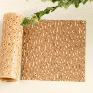 May include: A beige rolling pin and a flat, rectangular surface, both embossed with a repeating pattern of musical notes. The musical notes are raised and create a textured effect. The items are on a light background.