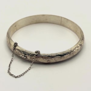 May include: A silver-colored bangle bracelet with an engraved floral pattern. The hinged bracelet has a safety chain and clasp closure. The jewelry is displayed on a white surface.