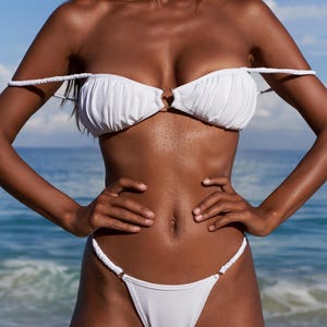 Könnte beinhalten: Weißes Bikini-Set mit gerüschtem Oberteil und String-Hose. Das Oberteil hat ein Ringdetail in der Mitte. Der Bikini ist aus einem glatten, einfarbigen weißen Stoff gefertigt. Der Hintergrund zeigt das Meer und einen blauen Himmel.