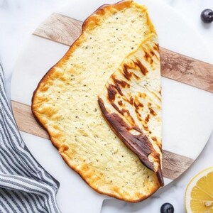 May include: A folded, golden-brown flatbread with a slightly charred edge, resting on a white and wood-grain cutting board. The bread has a textured surface with small, dark specks. A striped cloth and a lemon half are visible.