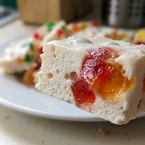 Puede incluir: Primer plano de una rebanada de postre blanco con trozos de fruta translúcidos y coloridos. El postre está en un plato blanco, con más rebanadas en el fondo. Los trozos de fruta son rojos, naranjas, verdes y amarillos.