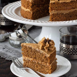 Könnte beinhalten: Ein Stück mehrschichtiger Kaffeekuchen mit einer Wirbelglasur und Kaffeebohnen auf einem weißen Teller. Der Kuchen steht auf einer weißen Kuchenplatte, mit einem ganzen Kuchen im Hintergrund. Ein Glas dunkler Flüssigkeit befindet sich in einem dekorativen Halter.