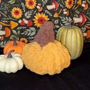 May include: A collection of decorative pumpkins including a small white pumpkin, a small orange pumpkin, a large textured yellow yarn pumpkin with a brown stem, and a large ribbed green pumpkin, all arranged against a dark floral fabric backdrop.