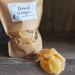 May include: A brown paper bag with a clear window containing dried ginger slices. The label reads "Dried Ginger" and "Herbal Haven". A small pile of ginger slices is in front of the bag on a wooden surface.
