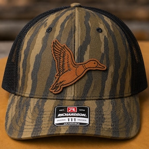 May include: A camouflage trucker hat with a black mesh back and a brown leather duck patch. The hat features the text "RICHARDSON 111 TRUCKER ADJUSTABLE" on a tag.