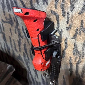 May include: A red and black multi-tool with the brand name "URBA" visible. The tool is secured with a black strap and mounted on a patterned surface. The tool includes a key ring and a black handle.