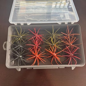 May include: A gray plastic storage box containing twelve multi-legged fishing lures. The lures are arranged in a grid and feature various colors including black, olive green, orange, and reddish-brown, each with numerous thin, flexible appendages.