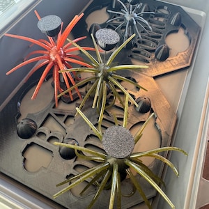 May include: Three multi-legged fishing lures with textured circular tops, one in orange and two in olive green, resting on a black 3D-printed plastic tray with cutouts and the embossed text DONKEY. The lures feature long, flexible, radiating tentacles.