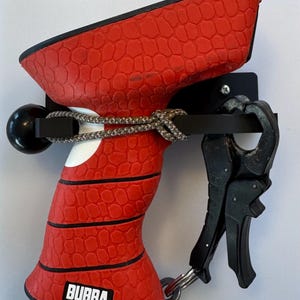 May include: A red and black spearfishing gun with a textured grip and a black metal hook. The gun is mounted on a black metal wall mount with a rope attached to the handle. The gun has the word "BUBBA" printed on the side.